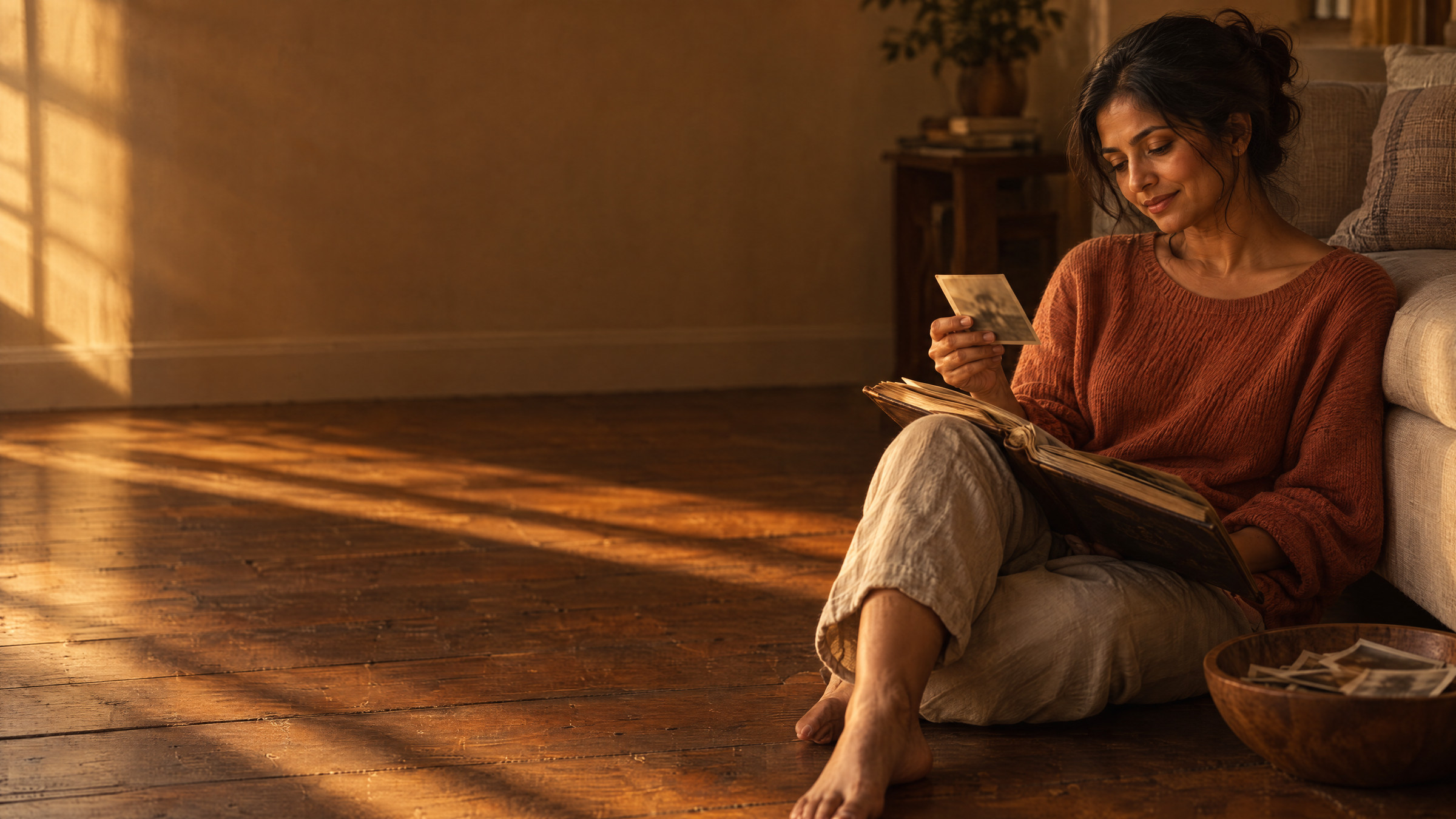 A woman sitting on a sunlit wooden floor with an old photo album open in her lap, studying one photograph