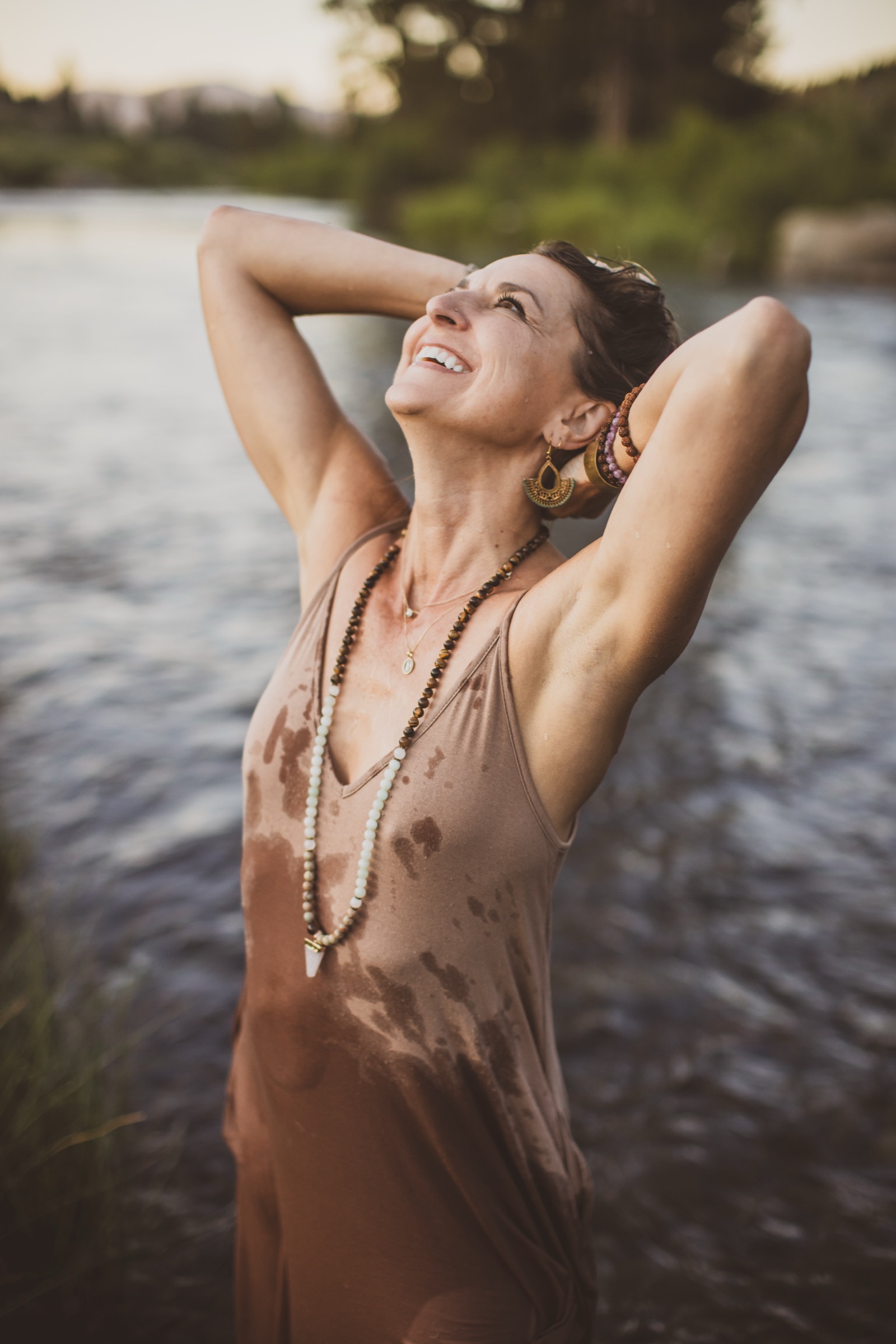 A woman by the river at golden hour, arms raised, laughing, mala beads catching the light