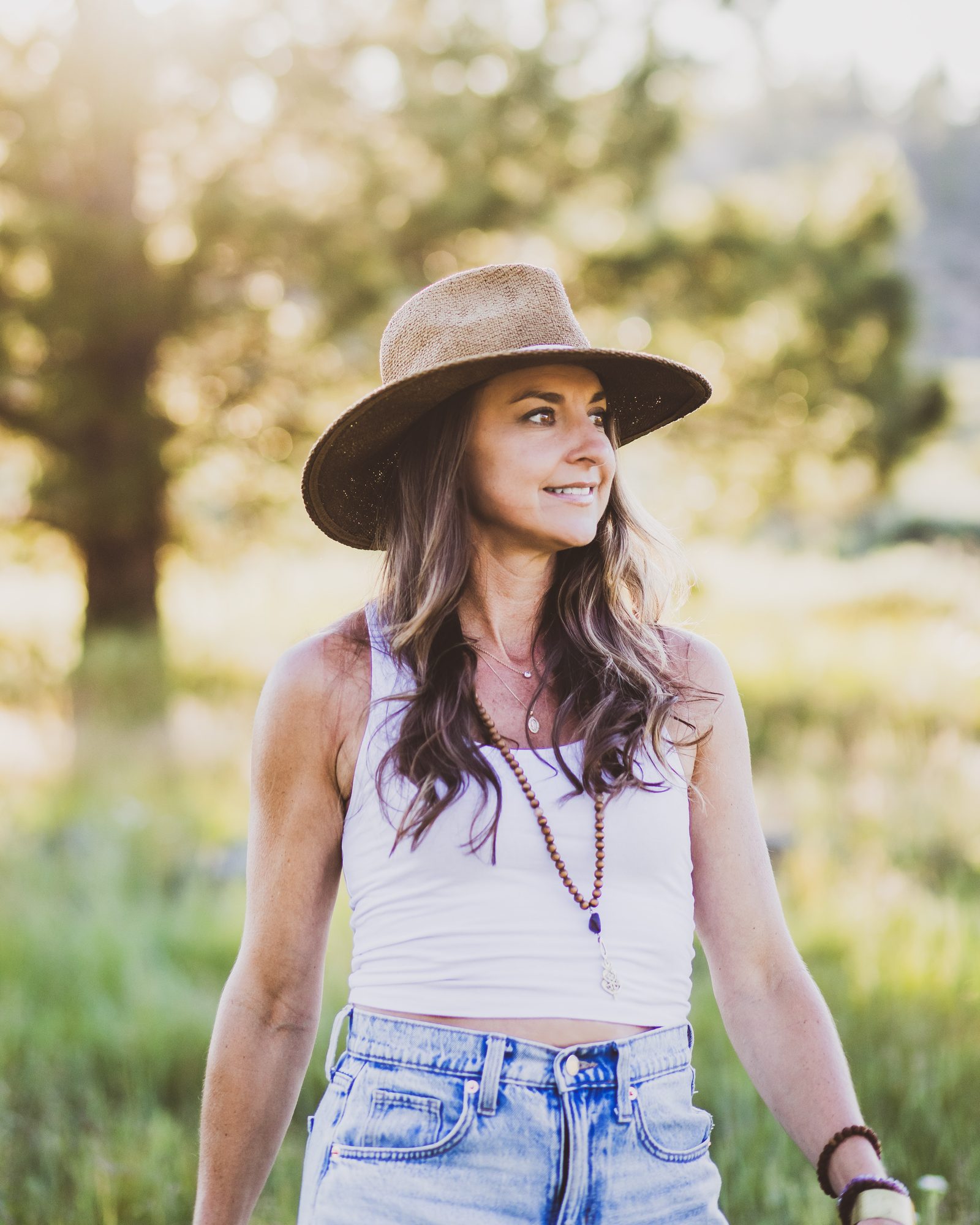 Kaila Leilani in a straw hat in a sunlit meadow, smiling and looking to the side, a long mala necklace over a white top, a tree glowing in the light behind her